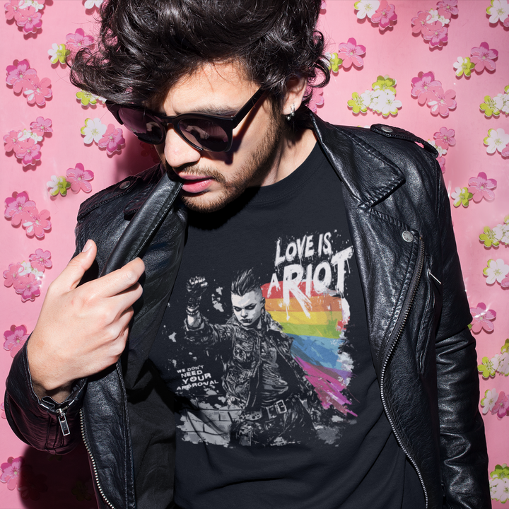 Model wearing a black Pride Riot t-shirt in an urban setting. The shirt features the phrase 'LOVE IS A RIOT' in distressed white punk-style lettering. High-contrast streetwear aesthetic.